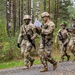 Four U.S. Army soldiers in MultiCam (OCP) camouflage uniforms are walking along a gravel path through a forested area, likely during a field training exercise. Two soldiers in the foreground are reviewing documents or maps while moving, and two soldiers in the background have foliage attached to their helmets as camouflage. All soldiers are armed with M4-style carbines and wear full combat gear.