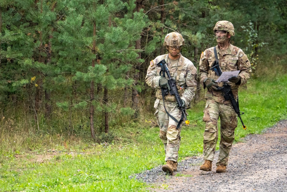 Two U.S. Army soldiers in Operational Camouflage Pattern (OCP) uniforms walk along a gravel path at the edge of a forested area. Both are carrying M4-style rifles and appear to be engaged in a field training exercise, with one soldier holding what appears to be a map or printed document. The setting suggests a military training environment, likely at a U.S. Army training installation.