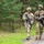7th Army NCOA Class 09-26 Land Nav Two U.S. Army soldiers in Operational Camouflage Pattern (OCP) uniforms walk along a gravel path at the edge of a forested area. Both are carrying M4-style rifles and appear to be engaged in a field training exercise, with one soldier holding what appears to be a map or printed document. The setting suggests a military training environment, likely at a U.S. Army training installation.