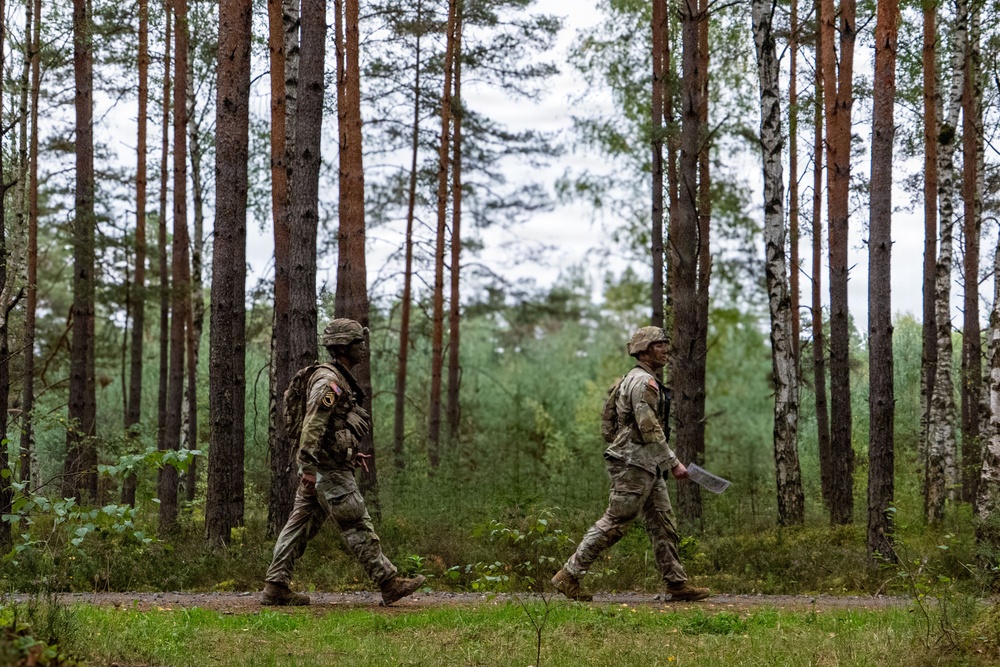 Two soldiers in camouflage uniforms are walking along a forest path among tall pine and birch trees. Both wear tactical gear and patches on their shoulders, with one soldier carrying what appears to be a document or map. The setting suggests a military training exercise or patrol in a northern European-style forest environment.