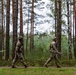 Two soldiers in camouflage uniforms are walking along a forest path among tall pine and birch trees. Both wear tactical gear and patches on their shoulders, with one soldier carrying what appears to be a document or map. The setting suggests a military training exercise or patrol in a northern European-style forest environment.