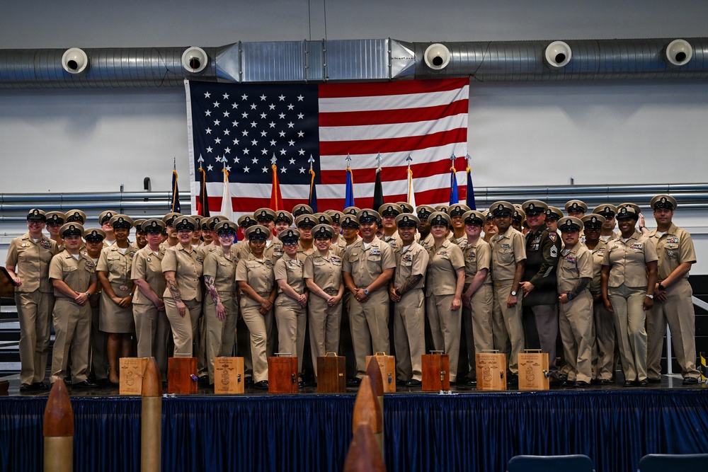 A large group of U.S. Navy personnel in khaki service uniforms pose for a formal group photograph on a stage. An American flag is prominently displayed behind them alongside multiple military unit flags. Wooden award plaques or boxes are arranged along the front of the stage, suggesting a recognition or graduation ceremony.