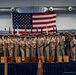 A large group of U.S. Navy personnel in khaki service uniforms pose for a formal group photograph on a stage. An American flag is prominently displayed behind them alongside multiple military unit flags. Wooden award plaques or boxes are arranged along the front of the stage, suggesting a recognition or graduation ceremony.