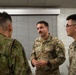 Three military personnel are engaged in conversation in what appears to be an indoor facility. Two U.S. Army soldiers in OCP (Operational Camouflage Pattern) uniforms are speaking with a third individual wearing a different camouflage pattern, likely from a foreign military. The interaction appears to be a professional or liaison meeting between allied forces.