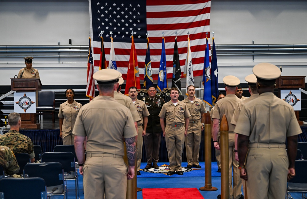 A formal U.S. military ceremony is taking place in an indoor venue, likely a change of command or awards ceremony. Personnel in Navy working uniforms (NWUs) and Army combat uniforms are assembled facing a stage area backed by a large American flag and multiple service branch flags. A speaker is at a podium on the left while a group of service members stands at attention on the stage.