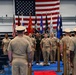A formal U.S. military ceremony is taking place in an indoor venue, likely a change of command or awards ceremony. Personnel in Navy working uniforms (NWUs) and Army combat uniforms are assembled facing a stage area backed by a large American flag and multiple service branch flags. A speaker is at a podium on the left while a group of service members stands at attention on the stage.