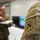 A close-up photograph of a U.S. Army soldier's shoulder sleeve insignia patch on an OCP (Operational Camouflage Pattern) uniform. The patch appears to be the U.S. Army Cyber Command or a related unit insignia featuring an arrow and stars in an oval design. In the background, two blurred soldiers are interacting near what appears to be a computer monitor in an indoor setting.