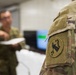 A close-up photograph of a U.S. Army soldier's shoulder sleeve insignia patch on an OCP (Operational Camouflage Pattern) uniform. The patch appears to be the U.S. Army Cyber Command or a related unit insignia featuring an arrow and stars in an oval design. In the background, two blurred soldiers are interacting near what appears to be a computer monitor in an indoor setting.