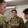 Three military personnel in camouflage uniforms are engaged in a discussion in what appears to be an indoor facility. The central figure, wearing glasses and holding a small notepad or booklet, appears to be communicating between the other two individuals. Two of the individuals wear a distinctive green camouflage pattern consistent with Japan Ground Self-Defense Force uniforms, while the figure on the left wears a different camouflage pattern.