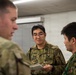 Three military personnel in camouflage uniforms are engaged in a discussion in what appears to be an indoor facility. The central figure, wearing glasses and holding a small notepad or booklet, appears to be communicating between the other two individuals. Two of the individuals wear a distinctive green camouflage pattern consistent with Japan Ground Self-Defense Force uniforms, while the figure on the left wears a different camouflage pattern.