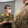 A Japan Ground Self-Defense Force (JGSDF) soldier, identified by name tape as 'A. NUMAKURA', wearing a white face mask and holding a notepad, appears to be in conversation with a U.S. Army soldier in MultiCam uniform. The setting appears to be an indoor military facility, suggesting a joint exercise, briefing, or coordination meeting between Japanese and American military personnel. A third soldier in MultiCam is visible in the background.