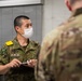 A Japan Ground Self-Defense Force (JGSDF) soldier, identified by name tape as 'A. NUMAKURA', wearing a white face mask and holding a notepad, appears to be in conversation with a U.S. Army soldier in MultiCam uniform. The setting appears to be an indoor military facility, suggesting a joint exercise, briefing, or coordination meeting between Japanese and American military personnel. A third soldier in MultiCam is visible in the background.