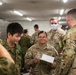 The image shows a joint military interaction between U.S. Army soldiers and what appear to be Japanese Ground Self-Defense Force (JGSDF) members, likely during a bilateral training exercise or exchange. A U.S. Army soldier named 'VEGA' is prominently centered, holding a notebook and smiling, appearing to facilitate communication or coordination. The setting is an indoor military facility with fluorescent lighting.