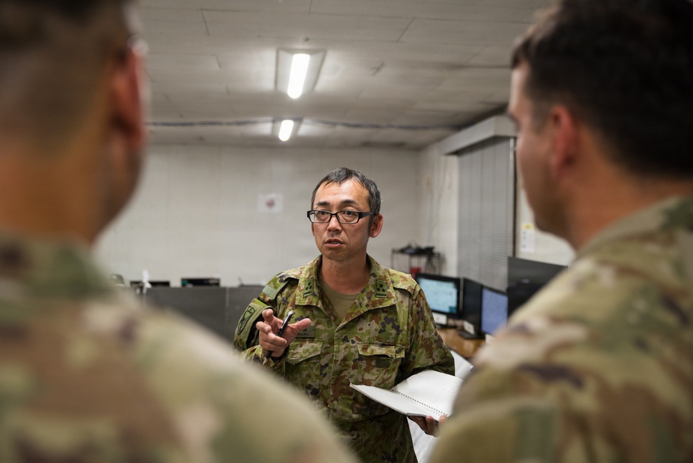 A military briefing or discussion is taking place indoors between at least three soldiers. The central figure, wearing a distinctive pixelated camouflage uniform consistent with Japan Ground Self-Defense Force (JGSDF) pattern, is speaking and holding a pen while another soldier holds a notepad. The setting appears to be a military operations or administrative room.