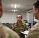A military briefing or discussion is taking place indoors between at least three soldiers. The central figure, wearing a distinctive pixelated camouflage uniform consistent with Japan Ground Self-Defense Force (JGSDF) pattern, is speaking and holding a pen while another soldier holds a notepad. The setting appears to be a military operations or administrative room.