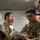 A group of military personnel in camouflage uniforms are gathered in what appears to be an indoor briefing or coordination session. Two distinct camouflage patterns are visible, suggesting personnel from different military forces are collaborating. The individuals are focused on documents and a tablet device, indicating a planning or information-sharing activity.