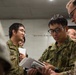 A group of military personnel in camouflage uniforms are gathered in what appears to be an indoor briefing or coordination session. Two distinct camouflage patterns are visible, suggesting personnel from different military forces are collaborating. The individuals are focused on documents and a tablet device, indicating a planning or information-sharing activity.