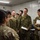 A group of military personnel in two distinct camouflage uniforms are gathered in what appears to be an indoor briefing or coordination room. The majority wear a green pixelated/woodland camouflage pattern consistent with Japan Ground Self-Defense Force (JGSDF) uniforms, while at least one individual on the left wears a MultiCam/OCP-style uniform. Several personnel are holding documents, clipboards, and papers, suggesting a planning or coordination meeting.