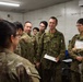 A group of military personnel in two distinct camouflage uniforms are gathered in what appears to be an indoor briefing or coordination room. The majority wear a green pixelated/woodland camouflage pattern consistent with Japan Ground Self-Defense Force (JGSDF) uniforms, while at least one individual on the left wears a MultiCam/OCP-style uniform. Several personnel are holding documents, clipboards, and papers, suggesting a planning or coordination meeting.