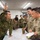 A joint military briefing or planning session between Japanese Ground Self-Defense Force (JGSDF) soldiers and U.S. Army personnel gathered around a tactical map on a table. A Japanese soldier is pointing upward while apparently explaining or briefing the group. U.S. Army soldiers are taking notes and listening attentively.