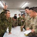 A joint military briefing or planning session between Japanese Ground Self-Defense Force (JGSDF) soldiers and U.S. Army personnel gathered around a tactical map on a table. A Japanese soldier is pointing upward while apparently explaining or briefing the group. U.S. Army soldiers are taking notes and listening attentively.