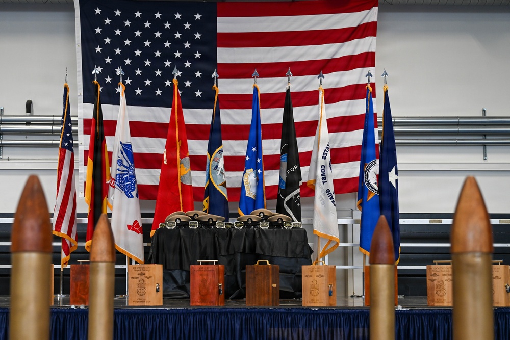 A formal military ceremony stage is displayed with a large American flag as a backdrop, flanked by multiple military branch and service flags. In the foreground, large artillery shell/bullet props are arranged in a row, while a draped table holds what appear to be awards, plaques, and military covers (hats). Wooden award boxes are visible along the base of the stage, suggesting a retirement, promotion, or awards ceremony.