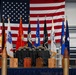 A formal military ceremony stage is displayed with a large American flag as a backdrop, flanked by multiple military branch and service flags. In the foreground, large artillery shell/bullet props are arranged in a row, while a draped table holds what appear to be awards, plaques, and military covers (hats). Wooden award boxes are visible along the base of the stage, suggesting a retirement, promotion, or awards ceremony.