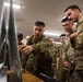 A group of military personnel in camouflage uniforms are gathered around a display screen or map board in what appears to be an indoor briefing or operations room. One soldier, wearing a distinctive green camouflage pattern consistent with Japanese Ground Self-Defense Force (JGSDF) uniform, is pointing at the display while others observe attentively. At least one soldier on the right wears a US Army MultiCam (OCP) uniform with a visible shoulder patch.