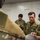 The image shows a group of military personnel in a briefing or coordination session. A U.S. soldier (visible from behind with an American flag patch on the left shoulder) is interacting with three soldiers wearing Japanese Ground Self-Defense Force (JGSDF) camouflage uniforms. The scene appears to be a joint military exercise or planning meeting taking place indoors.