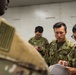 The image shows a group of military personnel in a briefing or coordination session. A U.S. soldier (visible from behind with an American flag patch on the left shoulder) is interacting with three soldiers wearing Japanese Ground Self-Defense Force (JGSDF) camouflage uniforms. The scene appears to be a joint military exercise or planning meeting taking place indoors.