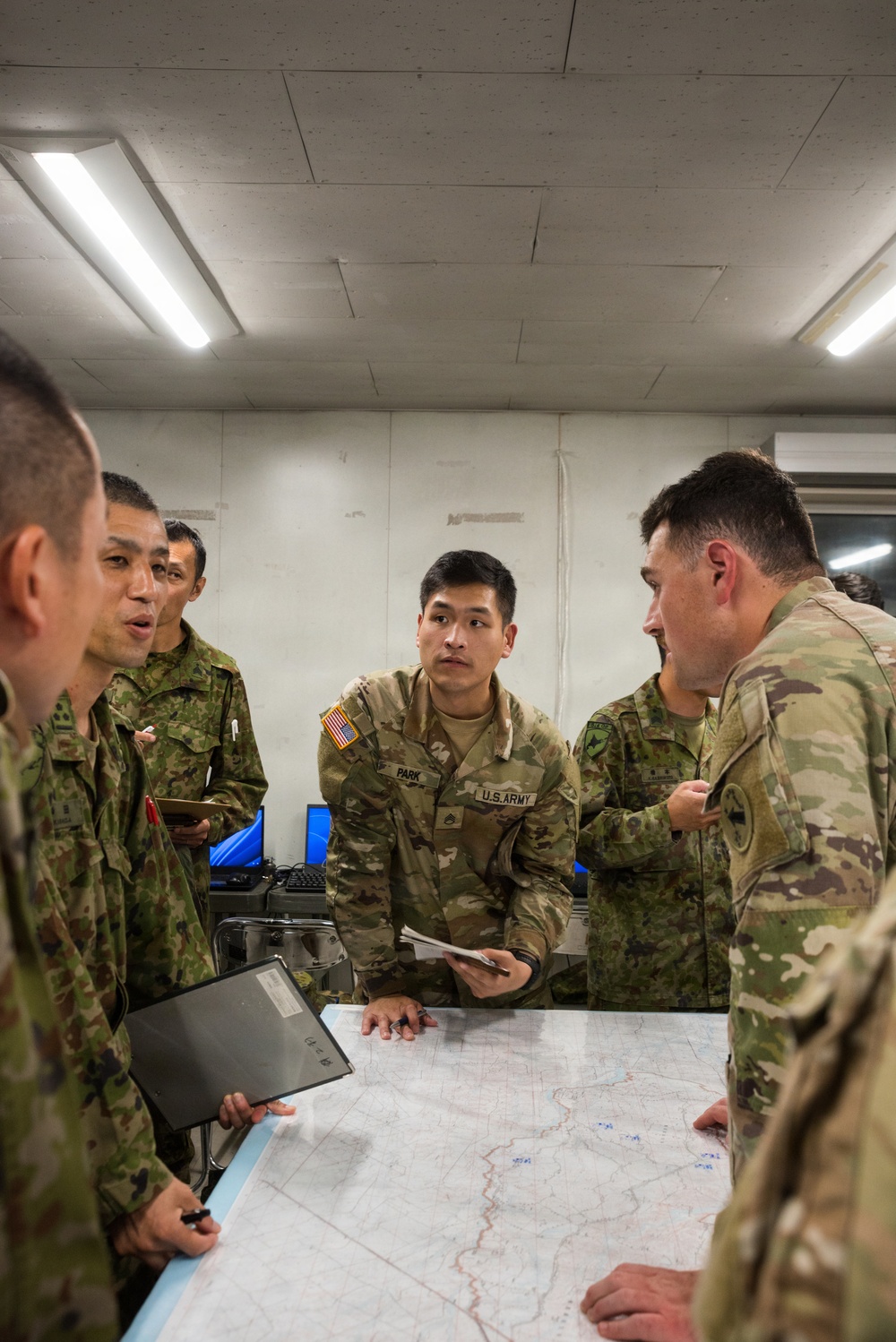 A group of military personnel from at least two different armed forces are gathered around a table with a map spread out, engaged in what appears to be a joint planning or briefing session. At least two soldiers are identifiable as U.S. Army personnel, while others wear a distinct camouflage pattern consistent with Japan Ground Self-Defense Force uniforms. The setting appears to be an indoor command or operations room with fluorescent lighting and computer equipment visible in the background.