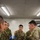 A group of military personnel from at least two different armed forces are gathered around a table with a map spread out, engaged in what appears to be a joint planning or briefing session. At least two soldiers are identifiable as U.S. Army personnel, while others wear a distinct camouflage pattern consistent with Japan Ground Self-Defense Force uniforms. The setting appears to be an indoor command or operations room with fluorescent lighting and computer equipment visible in the background.