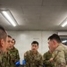 A group of military personnel from at least two different armed forces are gathered around a table with a map spread out, engaged in what appears to be a joint planning or briefing session. At least two soldiers are identifiable as U.S. Army personnel, while others wear a distinct camouflage pattern consistent with Japan Ground Self-Defense Force uniforms. The setting appears to be an indoor command or operations room with fluorescent lighting and computer equipment visible in the background.