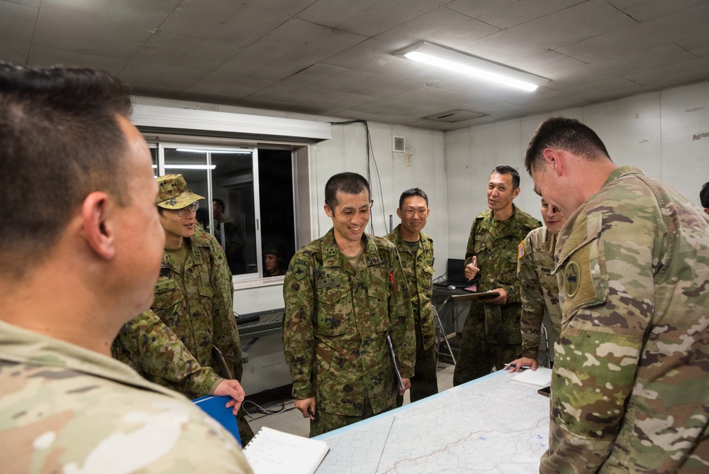 A group of military personnel from what appear to be two different armed forces are gathered around a table with maps spread out, engaged in a briefing or planning session. The soldiers on the left wear a distinctive green camouflage pattern consistent with Japan Ground Self-Defense Force uniforms, while the soldiers on the right wear OCP (Operational Camouflage Pattern) uniforms consistent with the U.S. Army. The setting appears to be an indoor command or operations room.