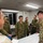 A group of military personnel from what appear to be two different armed forces are gathered around a table with maps spread out, engaged in a briefing or planning session. The soldiers on the left wear a distinctive green camouflage pattern consistent with Japan Ground Self-Defense Force uniforms, while the soldiers on the right wear OCP (Operational Camouflage Pattern) uniforms consistent with the U.S. Army. The setting appears to be an indoor command or operations room.