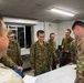 A group of military personnel from what appear to be two different armed forces are gathered around a table with maps spread out, engaged in a briefing or planning session. The soldiers on the left wear a distinctive green camouflage pattern consistent with Japan Ground Self-Defense Force uniforms, while the soldiers on the right wear OCP (Operational Camouflage Pattern) uniforms consistent with the U.S. Army. The setting appears to be an indoor command or operations room.