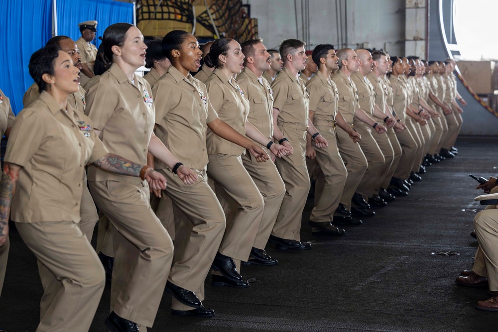 A large group of U.S. Navy personnel in tan/khaki service uniforms are performing what appears to be a coordinated ceremonial movement or chant, possibly a haka or similar group performance. They are lined up in a row inside what appears to be a ship's hangar bay, with all members in synchronized crouching stances with mouths open. The group is diverse in gender and ethnicity, all wearing matching Navy Working Uniforms.