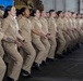 A large group of U.S. Navy personnel in tan/khaki service uniforms are performing what appears to be a coordinated ceremonial movement or chant, possibly a haka or similar group performance. They are lined up in a row inside what appears to be a ship's hangar bay, with all members in synchronized crouching stances with mouths open. The group is diverse in gender and ethnicity, all wearing matching Navy Working Uniforms.