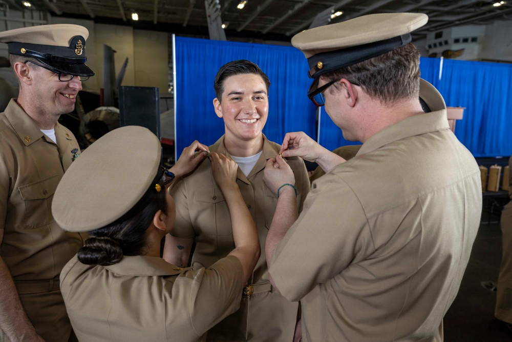A U.S. Navy promotion or pinning ceremony is taking place in what appears to be a ship's hangar bay. Two service members are simultaneously pinning insignia onto the collar of a smiling sailor in the center, while a third sailor looks on approvingly. The atmosphere is celebratory and the recipient appears proud and happy.
