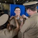 A U.S. Navy promotion or pinning ceremony is taking place in what appears to be a ship's hangar bay. Two service members are simultaneously pinning insignia onto the collar of a smiling sailor in the center, while a third sailor looks on approvingly. The atmosphere is celebratory and the recipient appears proud and happy.