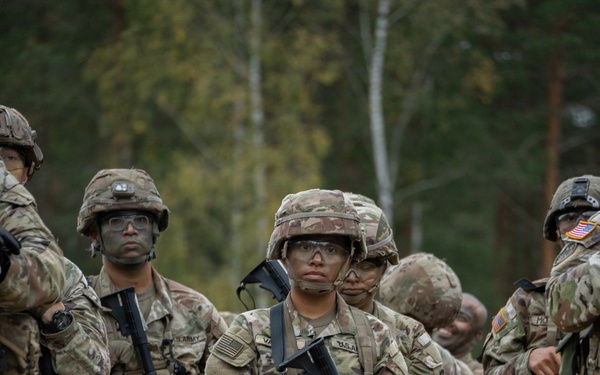 A group of U.S. Army soldiers in full combat gear are standing in formation in an outdoor wooded area. The soldiers are wearing Operational Camouflage Pattern (OCP) uniforms, helmets, and face camouflage paint, and are carrying M4/M16-style rifles equipped with yellow training device muzzle attachments. This appears to be a military training exercise based on the MILES/training gear indicators.