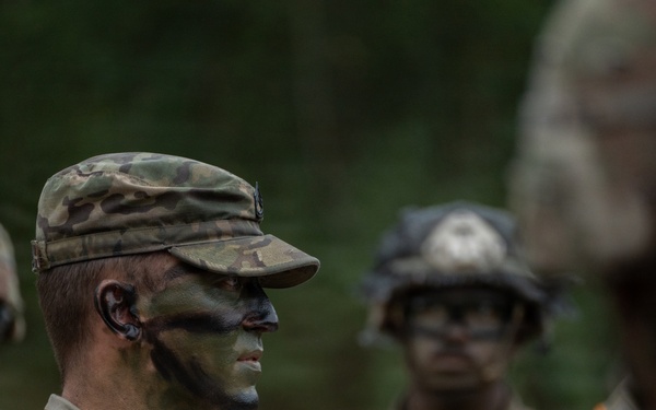 A group of military personnel in Operational Camouflage Pattern (OCP) uniforms are gathered outdoors, likely during a field training exercise. The central figure in sharp focus is wearing a patrol cap and has green and black camouflage face paint applied. Other soldiers visible in the background and foreground are wearing tactical helmets and protective eyewear.