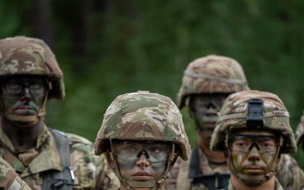 A group of U.S. Army soldiers in full combat gear, wearing Operational Camouflage Pattern (OCP) uniforms and helmets with face camouflage paint applied. The soldiers appear to be in a field training exercise, standing in formation in a wooded environment. The central soldier is holding a rifle and wearing protective eyewear, with a 'U.S. AR[MY]' tape visible on their uniform.