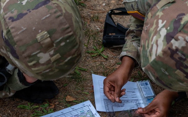 Two soldiers in US Army OCP (Operational Camouflage Pattern) uniforms are crouched on the ground conducting land navigation training. One soldier is holding a clear plastic protractor/coordinate scale over a printed topographic map laid on the ground. This appears to be a field training exercise focused on map reading and terrain navigation skills.