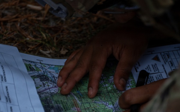 Military personnel in camouflage uniforms are conducting land navigation using a topographic map and what appears to be a compass or writing instrument. The individuals are positioned on the ground in an outdoor wooded environment, studying the map alongside a printed document that appears to be a grid reference or coordinate sheet. This is consistent with military field training exercises involving map reading and navigation skills.
