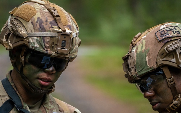 Two U.S. Army soldiers in full combat gear are reviewing a document or field reference card together in an outdoor wooded environment. Both wear OCP (Operational Camouflage Pattern) uniforms and FAST-style helmets with tactical accessories. The soldier on the right has a visible name tape reading 'MURILLO' and additional helmet markings including 'SPC' rank and blood type indicators.