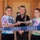 Three individuals are participating in a USO ribbon-cutting ceremony in front of what appears to be a temporary or forward-deployed facility. The center person holds scissors while all three grip a blue USO-branded ribbon. Two of the participants wear tie-dye USO t-shirts while the center person wears a black USO polo shirt.