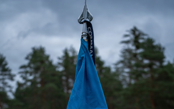 A blue military guidon flag bearing the 'NCOA' (Non-Commissioned Officer Academy) insignia is mounted on a wooden staff with a metal spear tip. The flag displays embroidered gold lettering and a patch featuring a triangular red and gold emblem. The setting appears to be an outdoor military training area with trees, overcast skies, and military vehicles visible in the background.