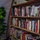 A wooden ladder-style bookshelf filled with numerous fiction and non-fiction books is displayed in what appears to be a cozy reading nook or glamping tent. The shelves contain a diverse collection of popular authors including David Baldacci, James Patterson, Bill Bryson, Greg Iles, and David Grann, among others. A fiddle-leaf fig plant is visible to the left, and a small potted green plant sits atop the bookshelf.