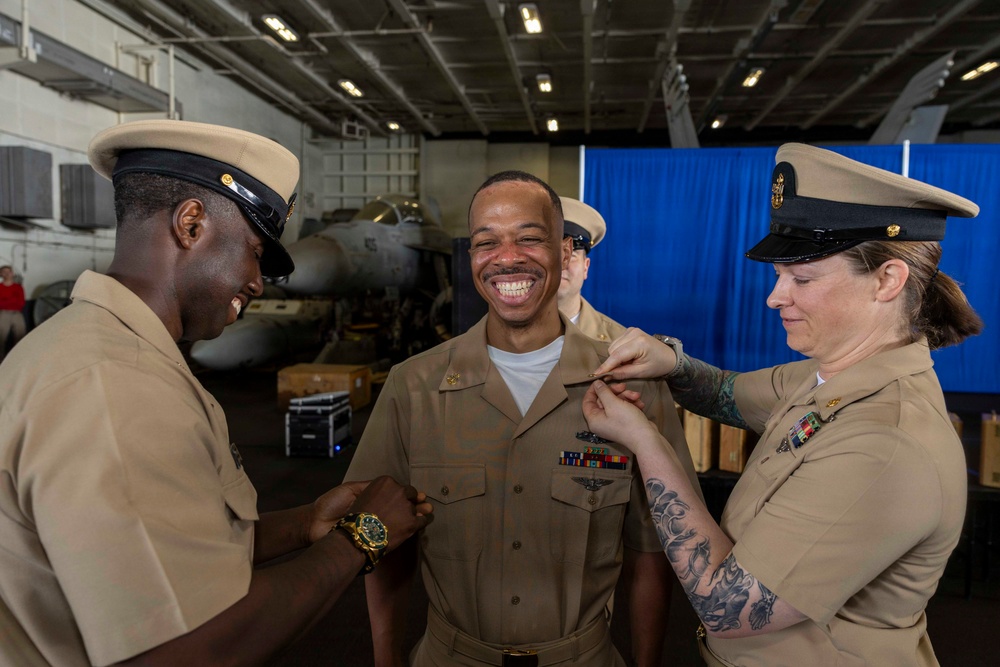 Three U.S. Navy personnel in khaki service uniforms are participating in what appears to be a pinning ceremony inside a ship's hangar bay. The central figure is smiling broadly while two colleagues — one on each side — pin insignia or devices onto his collar or shoulders. A military aircraft and blue ceremonial backdrop are visible in the background.